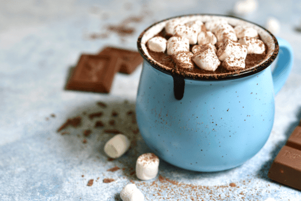 A blue mug of hot chocolate topped with marshmallows and cocoa powder, with chocolate pieces nearby.