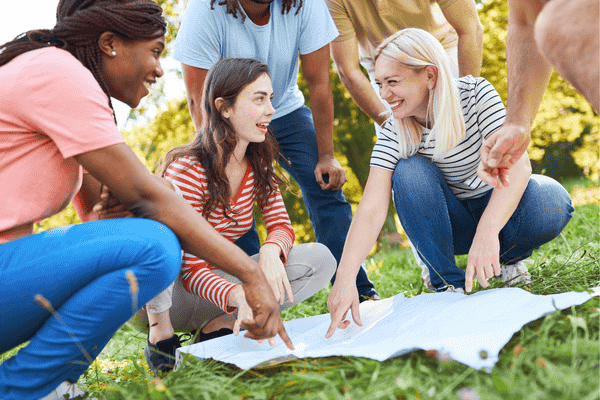 A group of people kneel on grass, smiling and pointing at a map spread out in front of them during a Family Photo Scavenger Hunt.