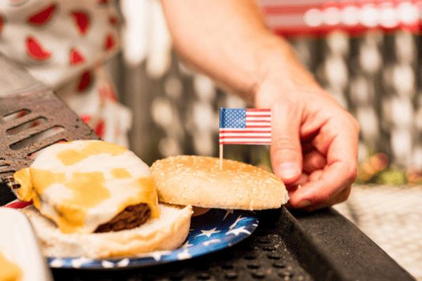 A hand holds a sesame bun next to a cheeseburger with an American flag toothpick on a blue star plate.