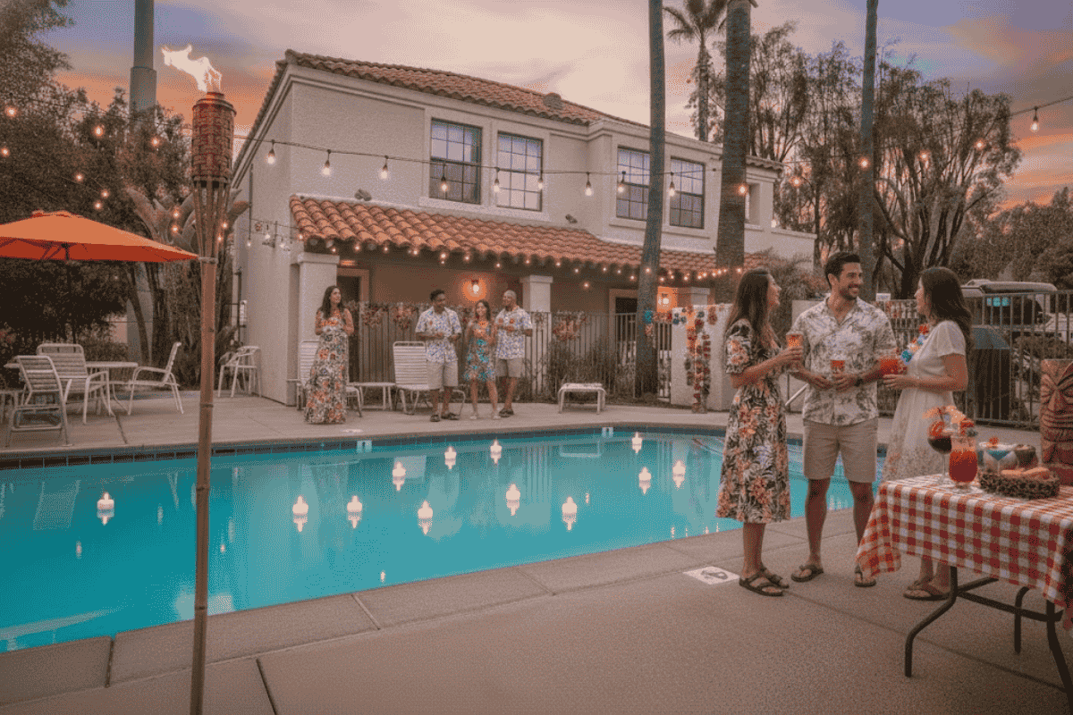 People in summer outfits gather around a pool at sunset, chatting near a table with drinks and snacks.