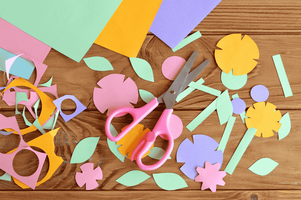 Pink scissors and colorful paper shapes for a craft project on a wooden table.