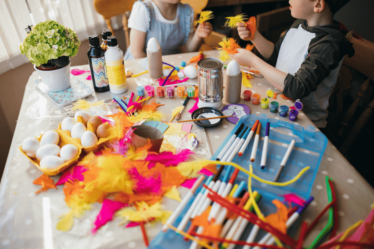 Two children doing arts and crafts with eggs, paints, feathers, brushes, and colorful materials on a table.