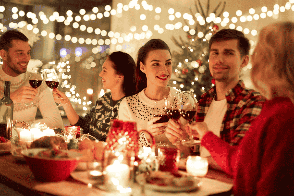 Five people celebrate at a festive dinner table, clinking glasses, with holiday lights and a Christmas tree behind them.