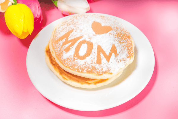 Two pancakes on a white plate with MOM and a heart dusted in powdered sugar, next to colorful tulips.