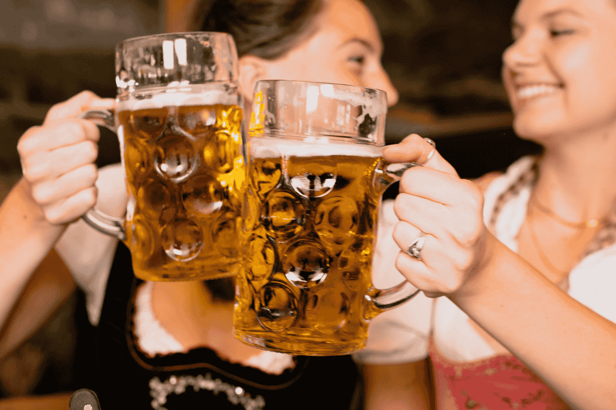 Two women in traditional dresses clink large beer mugs, smiling and celebrating together.
