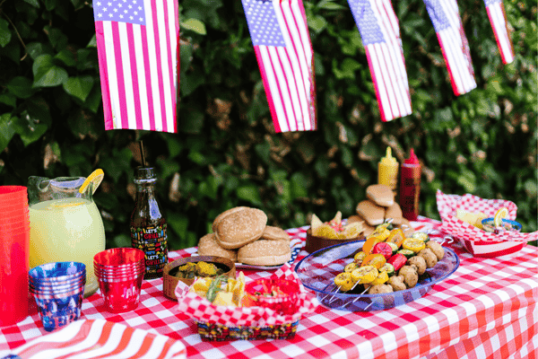 Festive outdoor picnic setup featuring a red and white checkered tablecloth, American flags, and a variety of foods including burgers, skewers, and colorful drinks, perfect for a summer gathering or Independence Day celebration.
