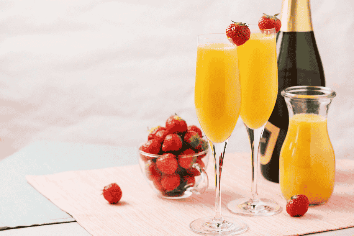 Two mimosas with strawberries, a glass pitcher of orange juice, and a bowl of fresh strawberries on a pink tablecloth.