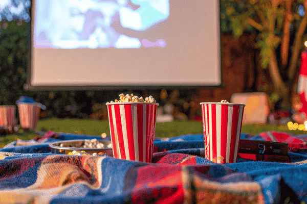 Outdoor movie night setup featuring two striped popcorn buckets on a colorful blanket, with a projection screen displaying a film in the background.