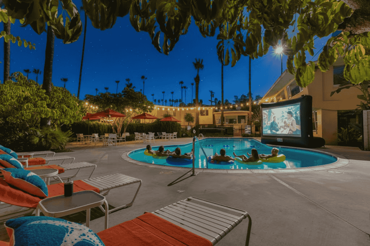 Outdoor movie night by the pool featuring guests relaxing on inflatable floats, surrounded by tropical plants and illuminated by string lights and a starry sky.