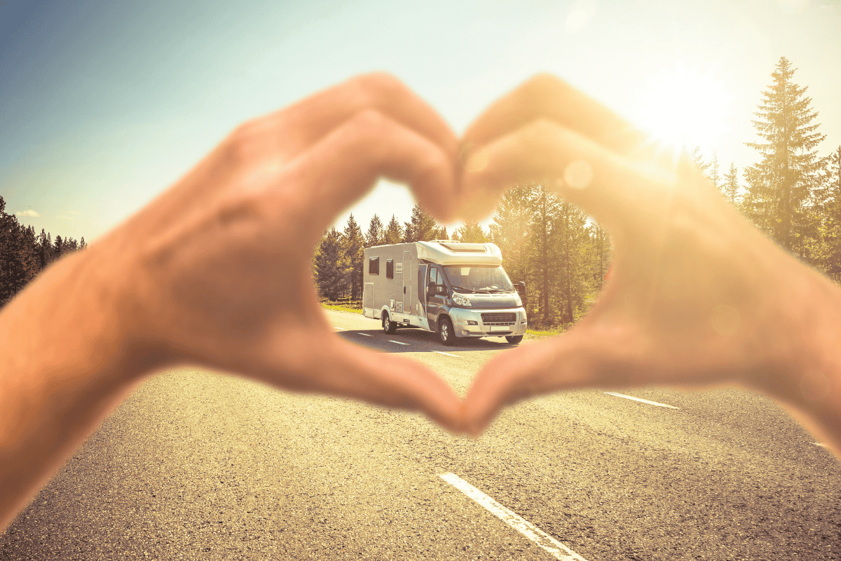 Hands forming a heart shape framing a motorhome on a sunny forest road
