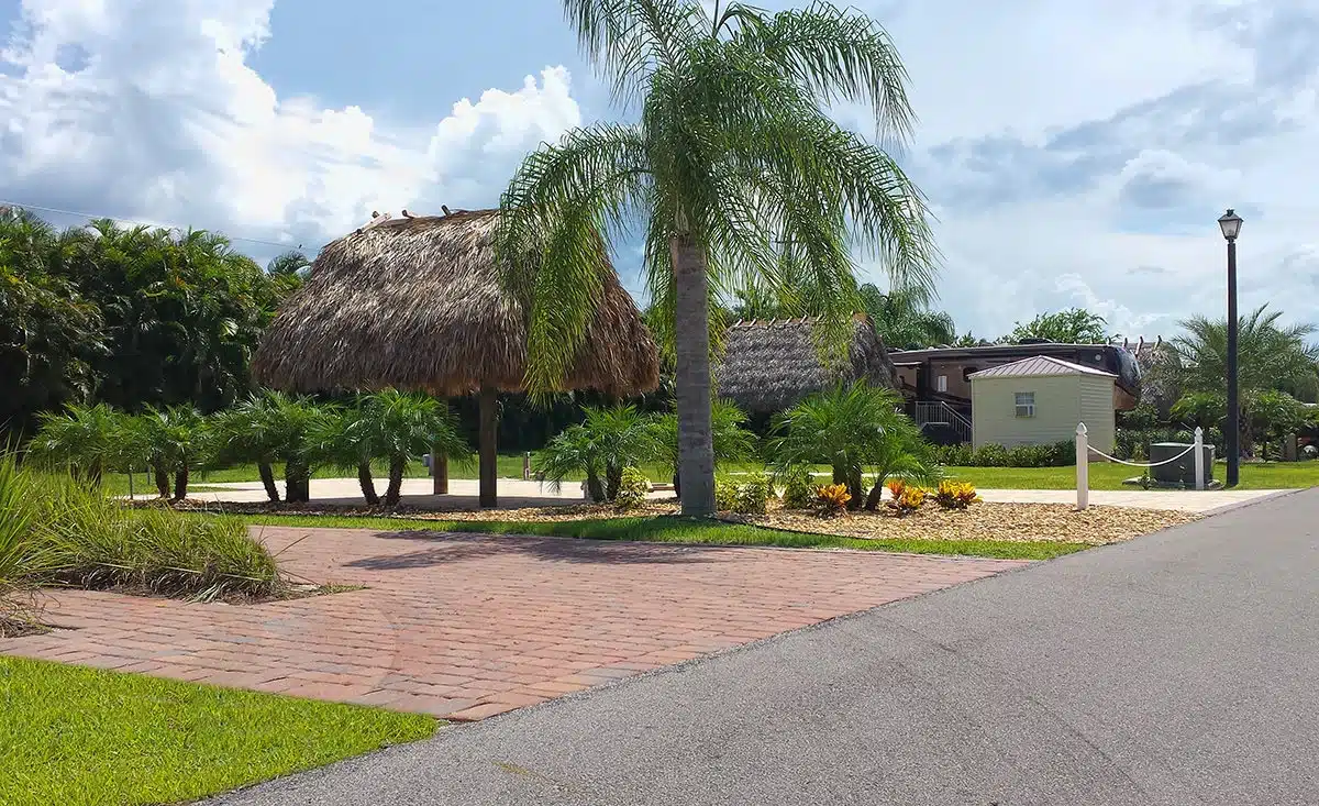 Palm trees and thatched huts line a paved area next to a road under a partly cloudy sky at Silver Palms RV Resort.